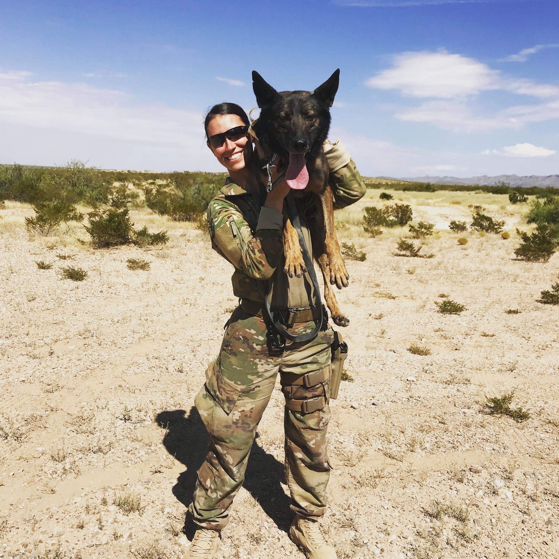 A woman in military uniform is holding a dog in the desert