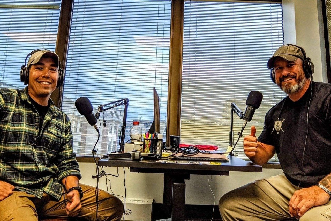 Two men are sitting at a table with microphones and giving a thumbs up.