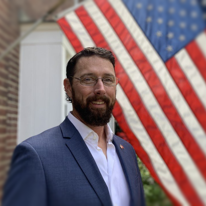 A man in a suit stands in front of an american flag