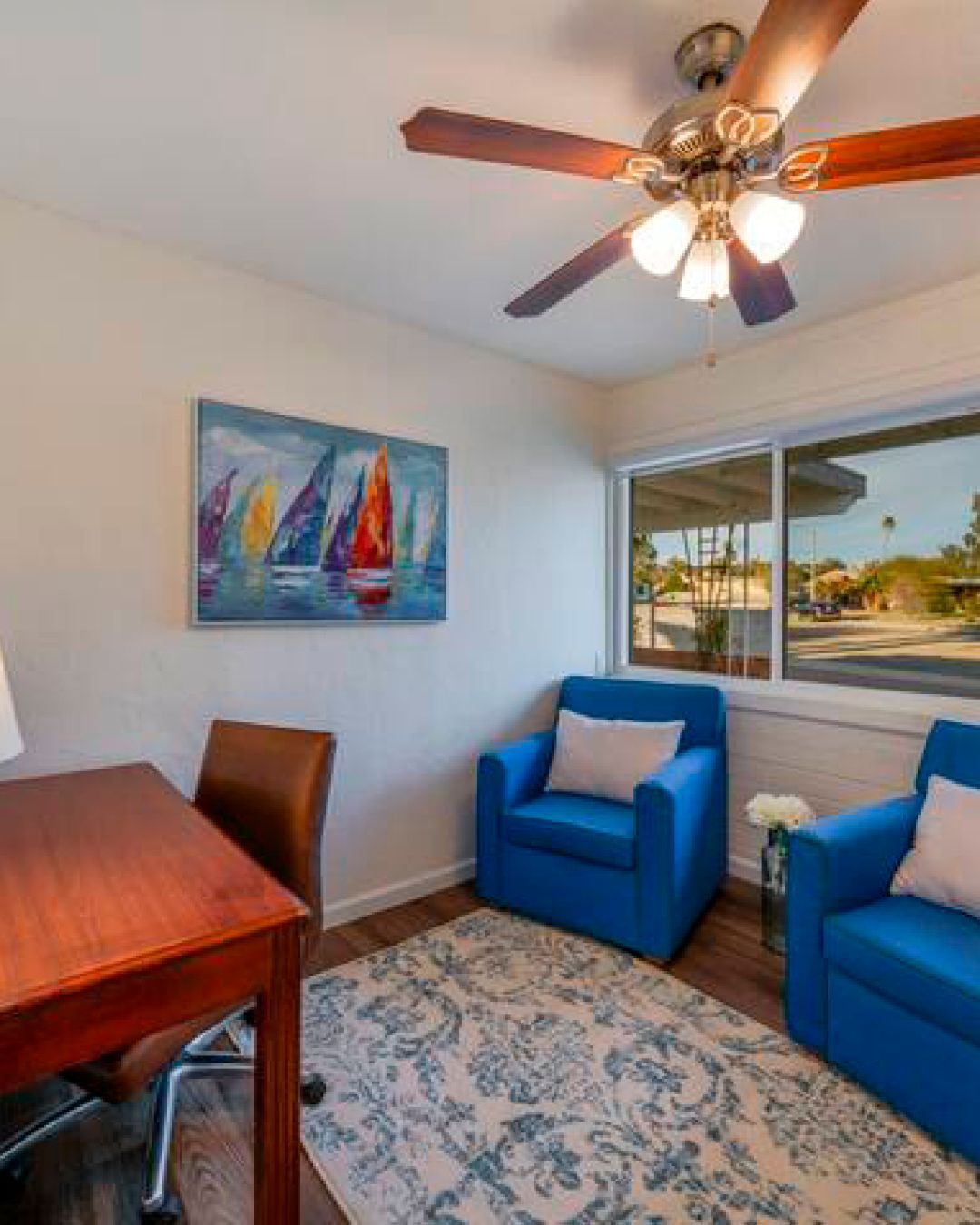 Bright office corner with desk, blue chairs, sailboat painting, patterned rug, and ceiling fan by a window