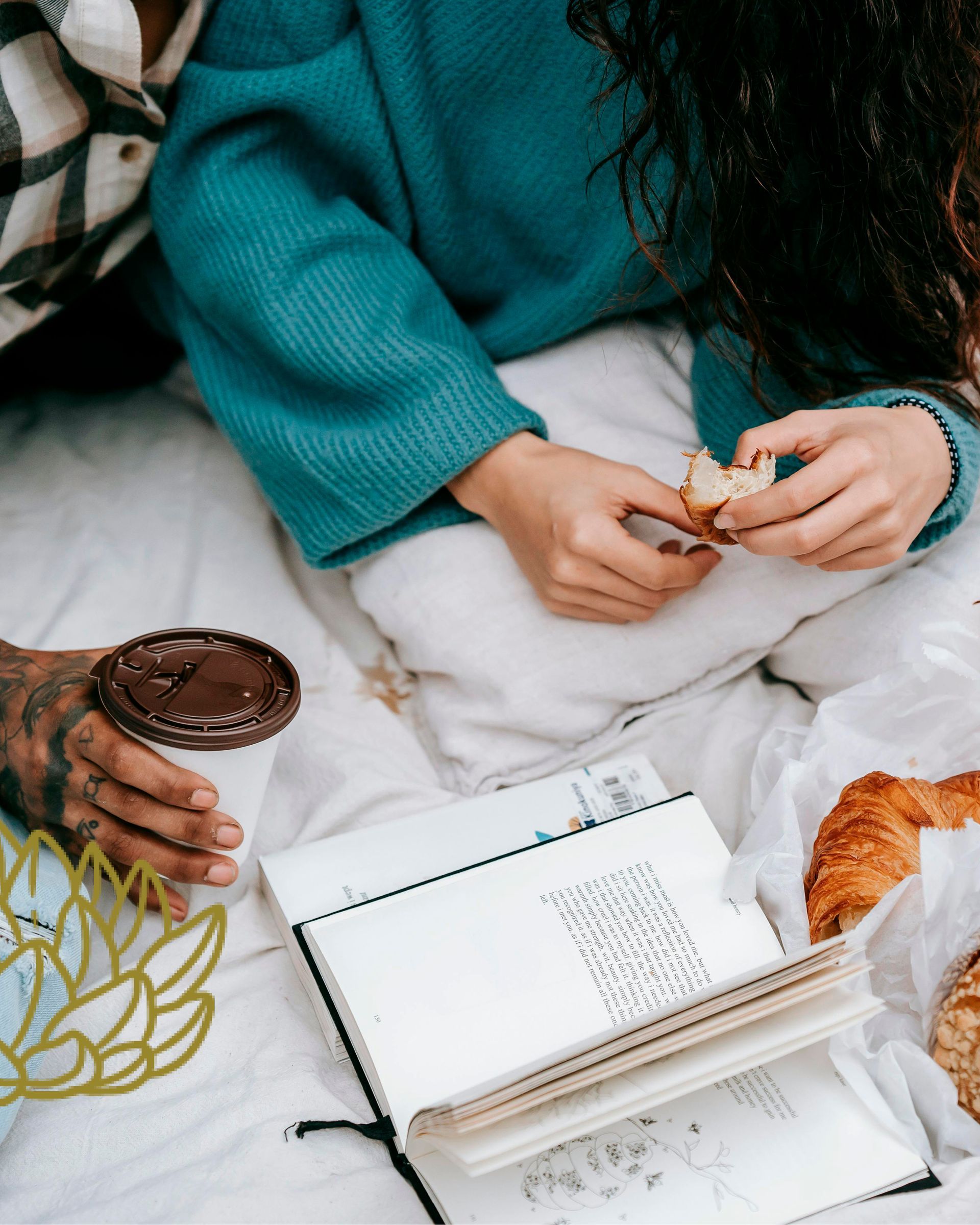 Person in teal sweater holding snack beside coffee, notebook, and croissant on a white blanket.