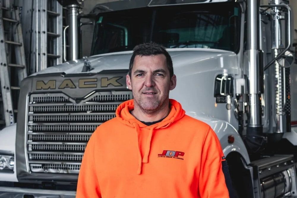 A Man In An Orange Hoodie Is Standing In Front Of A Mack Truck — Justin Drew Plumbing in Grovedale, VIC