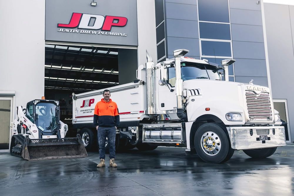 A Man Is Standing In Front Of A Dump Truck And A Bulldozer — Justin Drew Plumbing in Grovedale, VIC