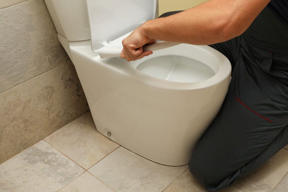 A Man Is Kneeling Down To Fix A Toilet In A Bathroom — Justin Drew Plumbing in Grovedale, VIC