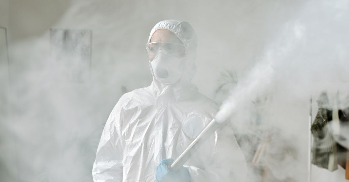 Una persona con traje protector y careta facial rocía desinfectante en un área interior con niebla.