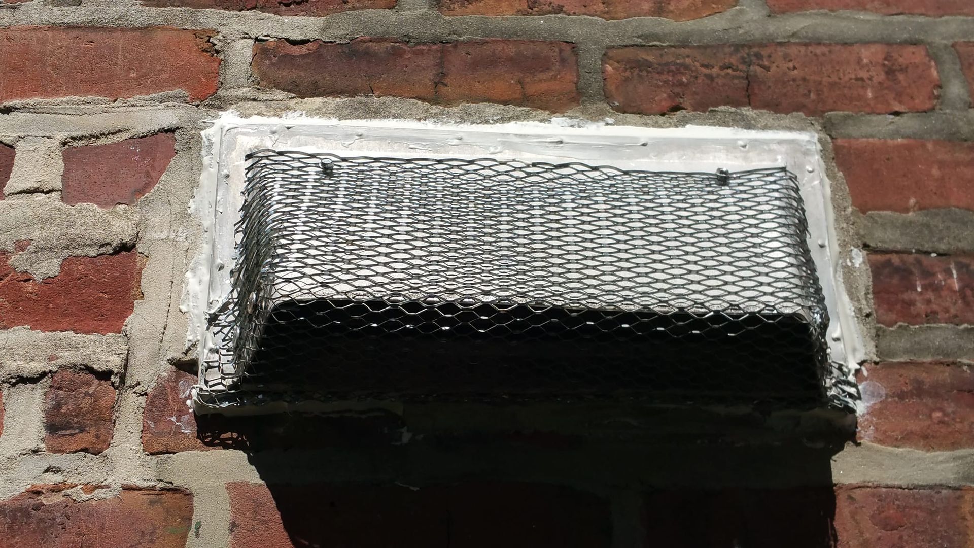 Brick wall with a rectangular vent covered by wire mesh, framed in gray concrete.