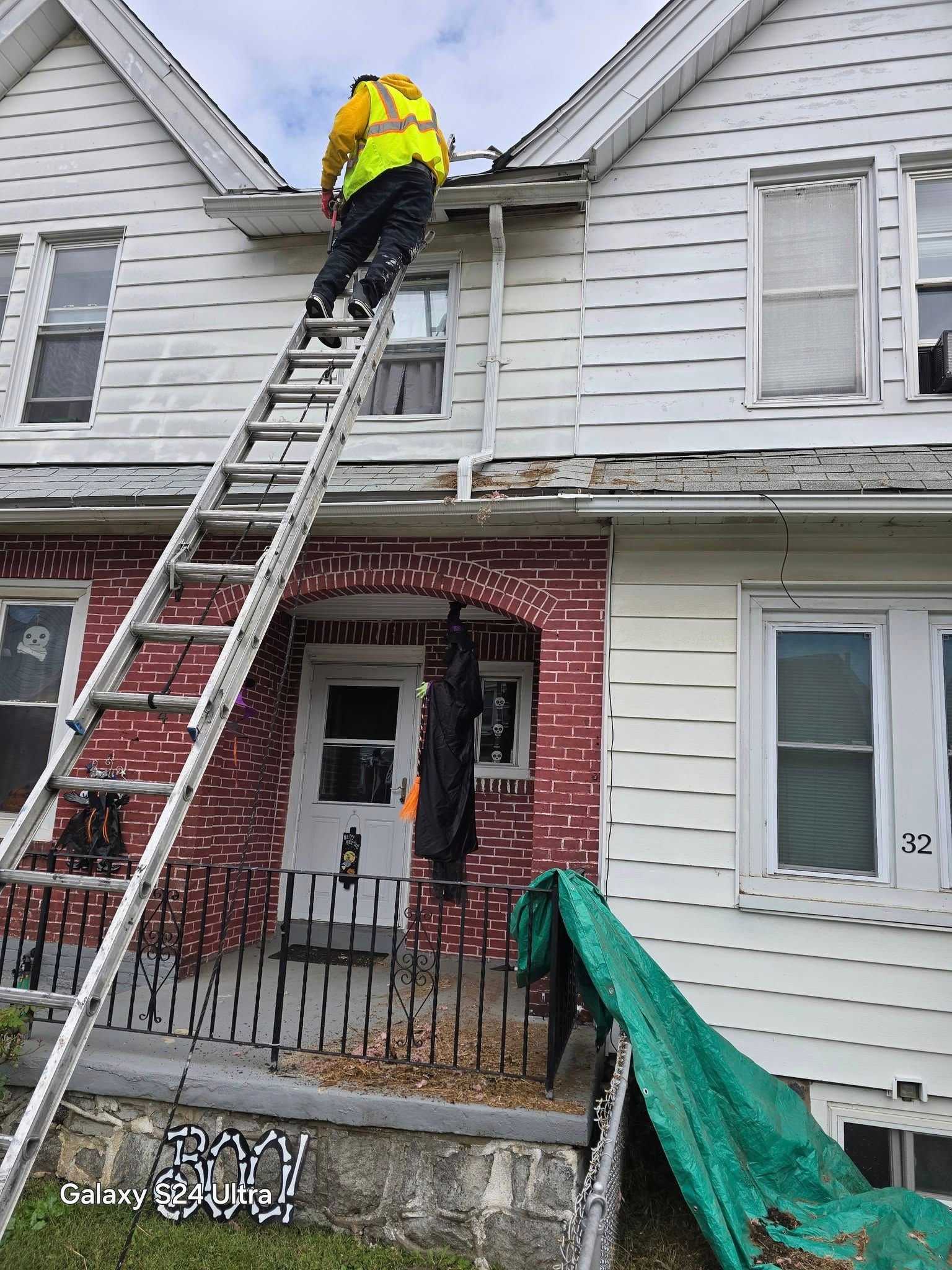 Person on a ladder, working on a two-story house with red brick and peeling white paint, wearing a yellow safety vest.