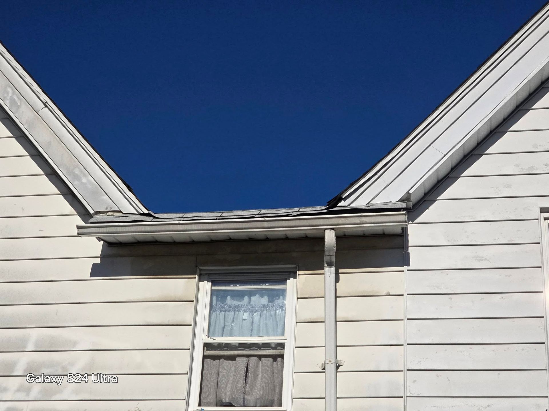 Exterior view of a weathered white house with a visible window under a clear, blue sky.