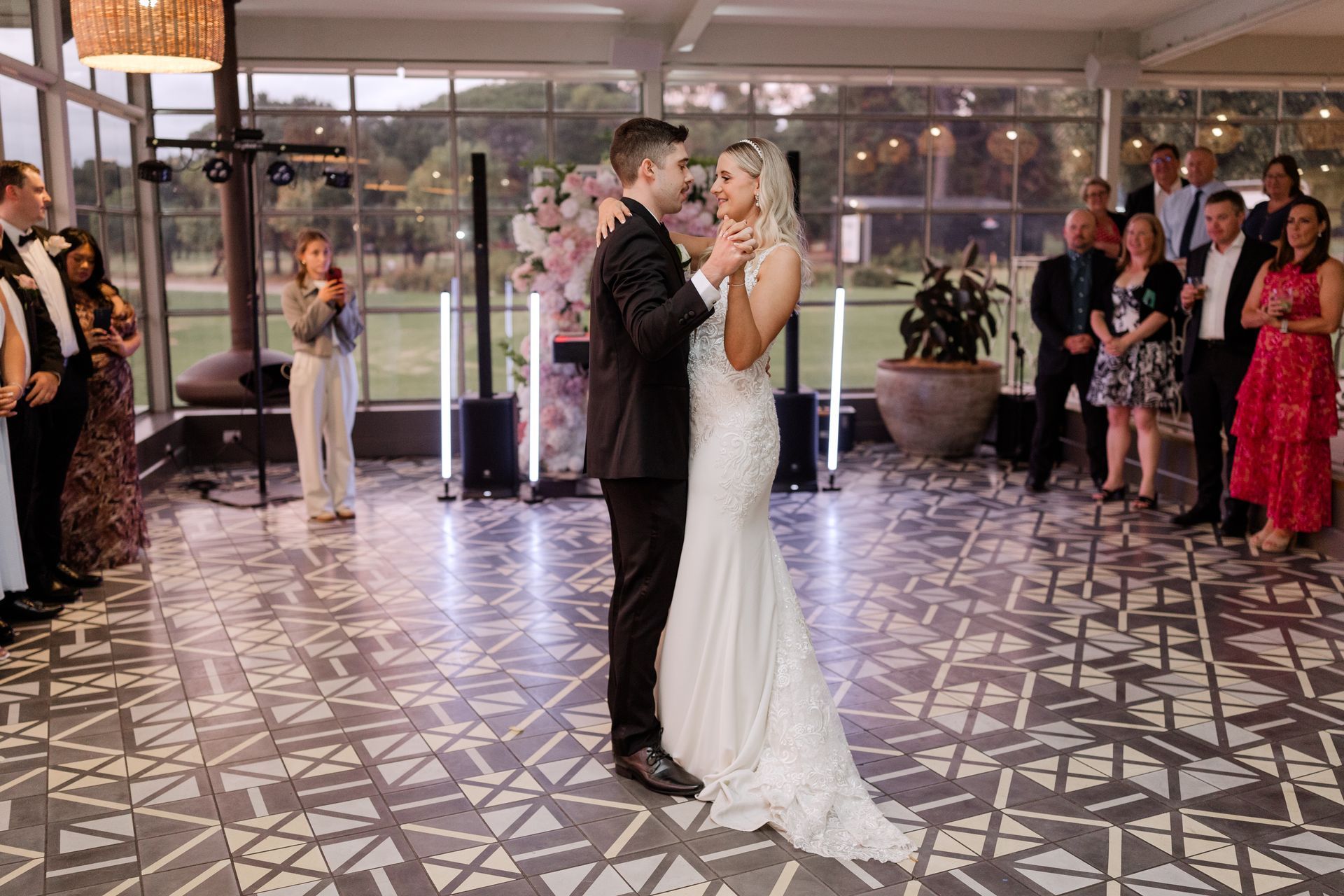 Couple dancing at a wedding reception, surrounded by guests. The bride wears a white gown, the groom a dark suit.