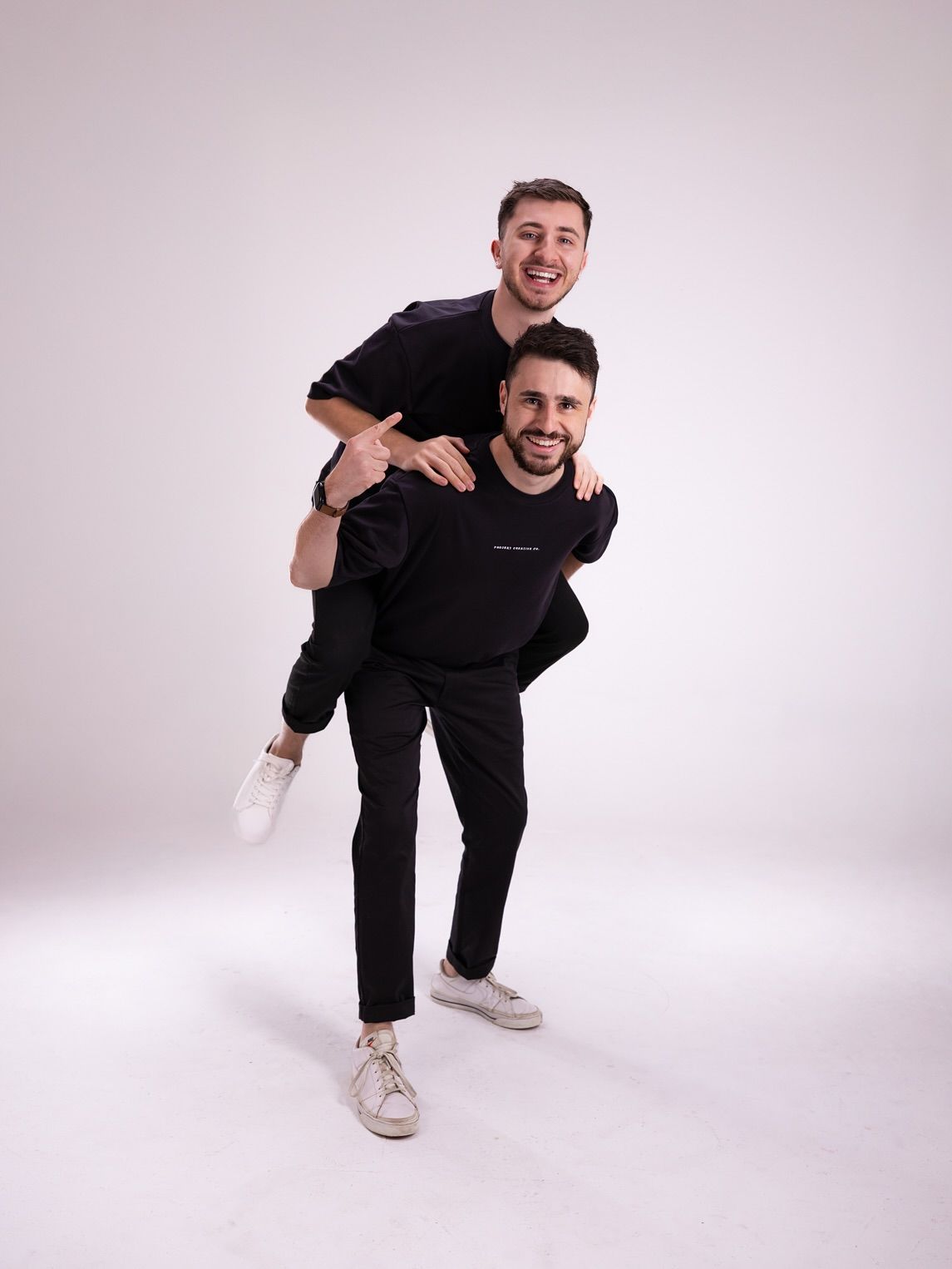 Two men wearing black clothing, one giving the other a piggyback ride, smiling, against a white background.
