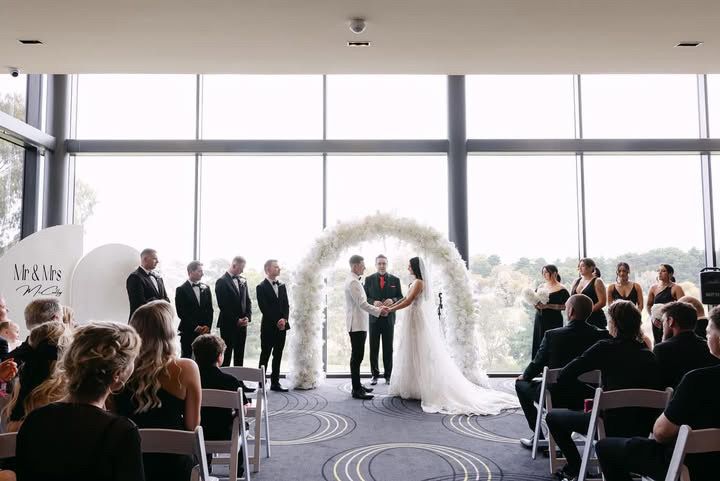 Wedding ceremony with couple under floral arch, window backdrop, guests seated.