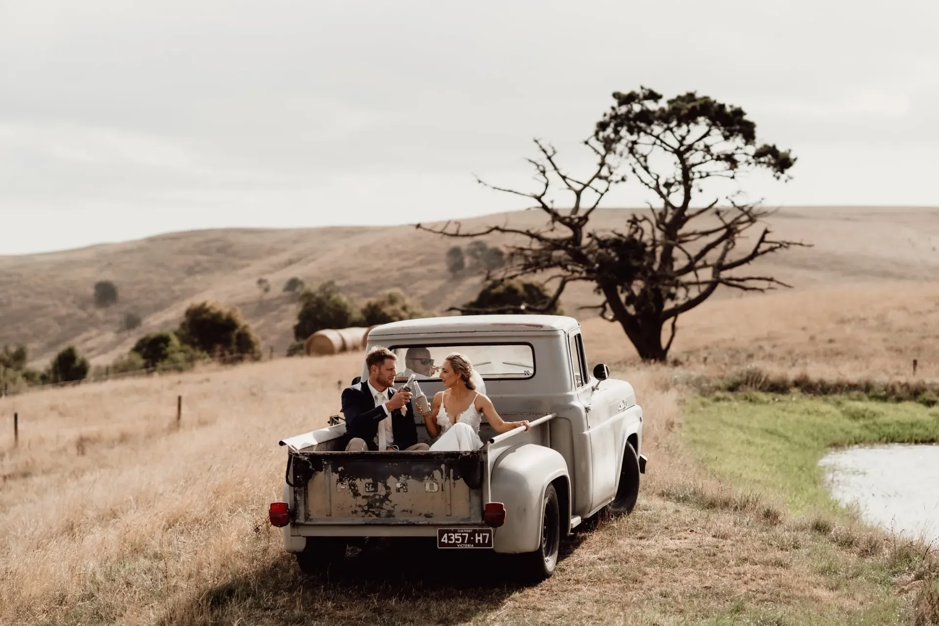 Newlyweds in a vintage truck, smiling, drinking champagne, beside a pond and rolling hills.