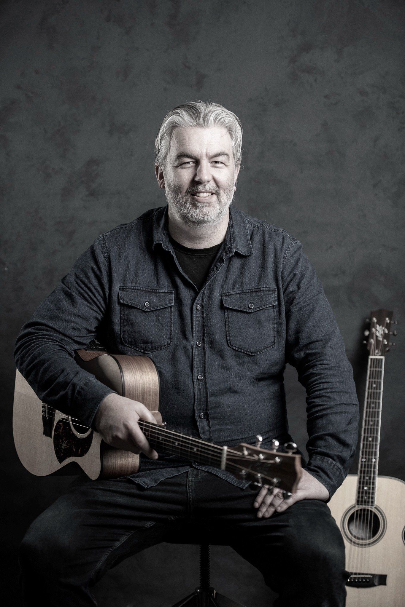 A man with a beard is sitting on a stool holding an acoustic guitar.