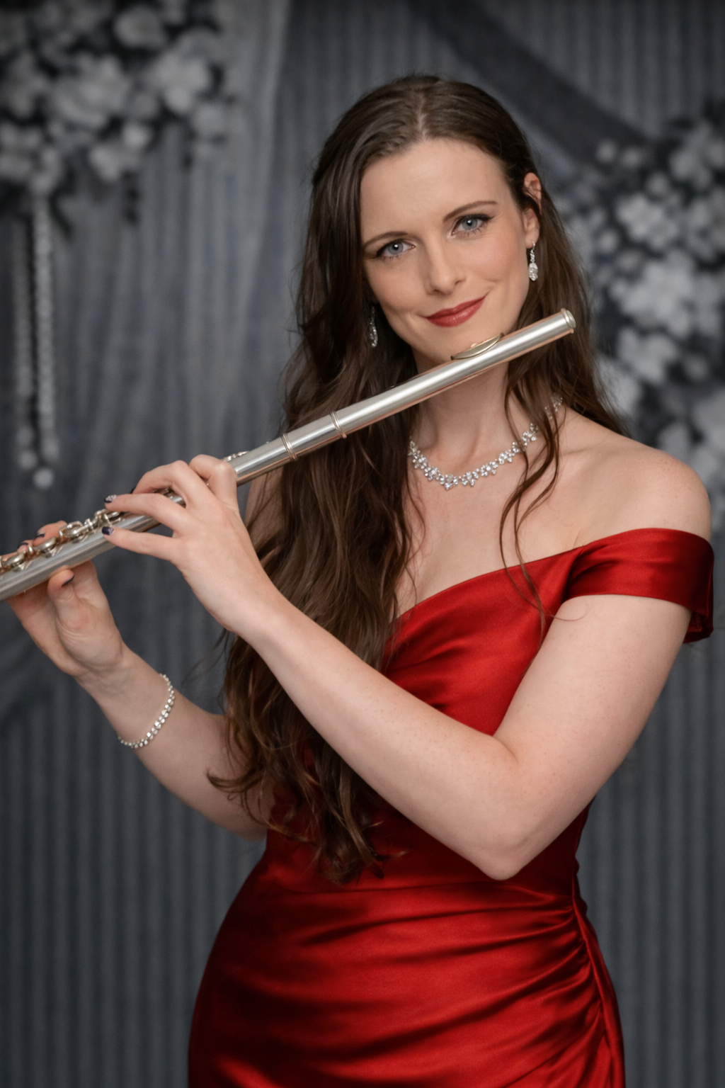 Woman in red off-shoulder dress plays a silver flute, smiling against a gray backdrop with floral accents.