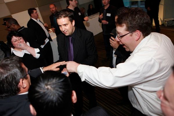 People interacting and laughing at a gathering. One man in a suit points, another in a white shirt smiles.