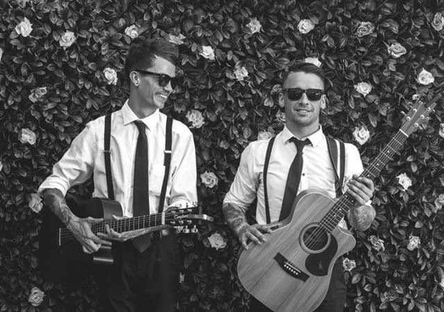 Two men are standing next to each other holding guitars in front of a wall of flowers.