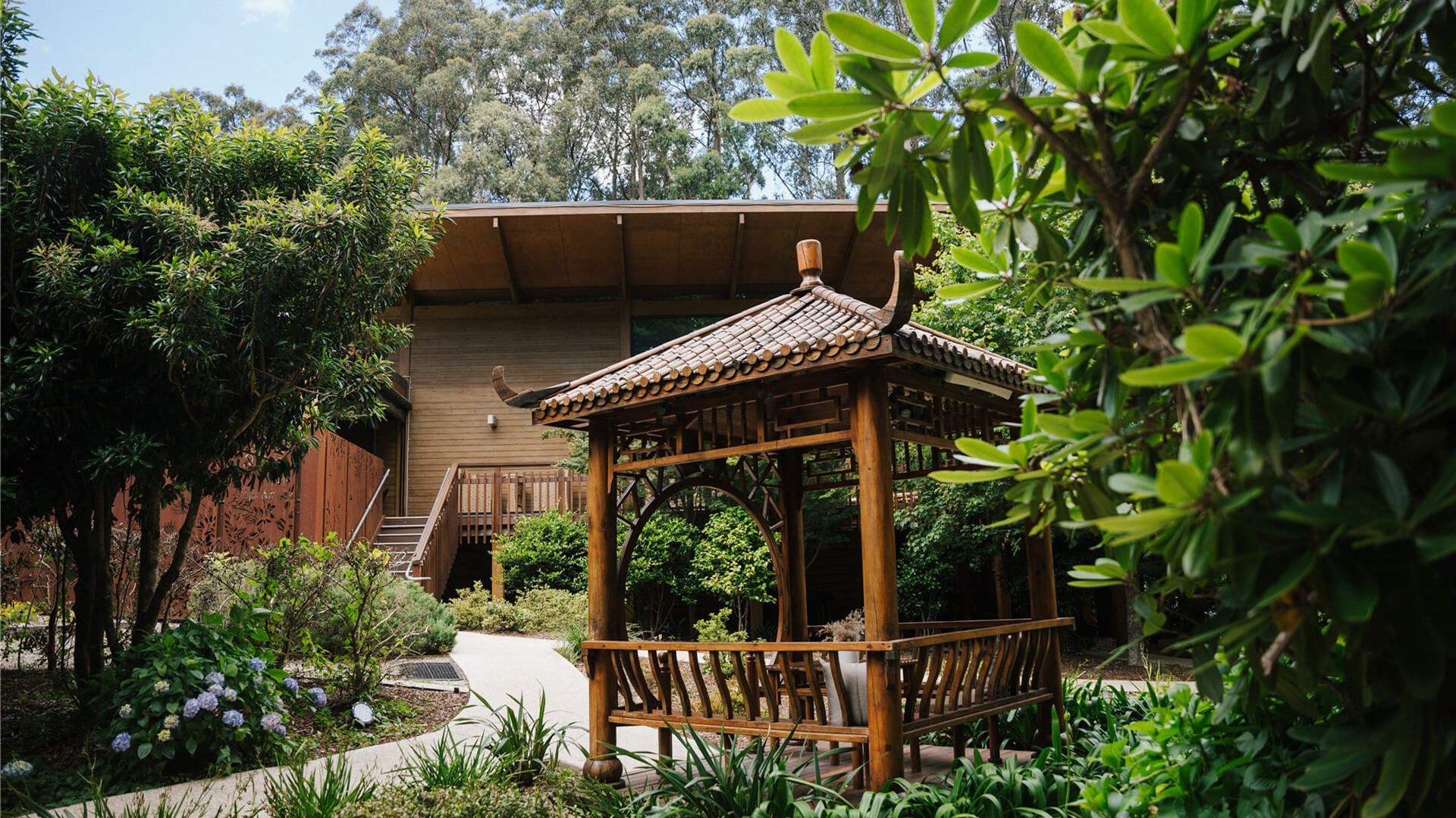 A wooden gazebo in a garden in front of a house surrounded by trees.