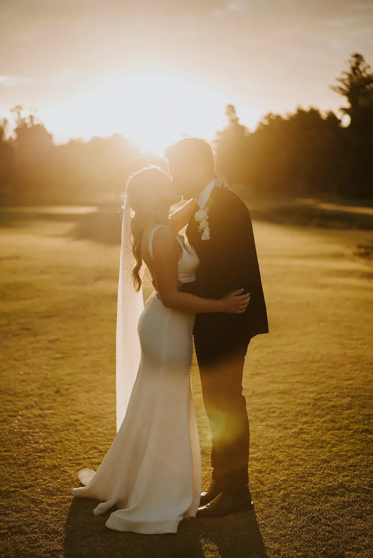 A bride and groom are kissing in a field at sunset.