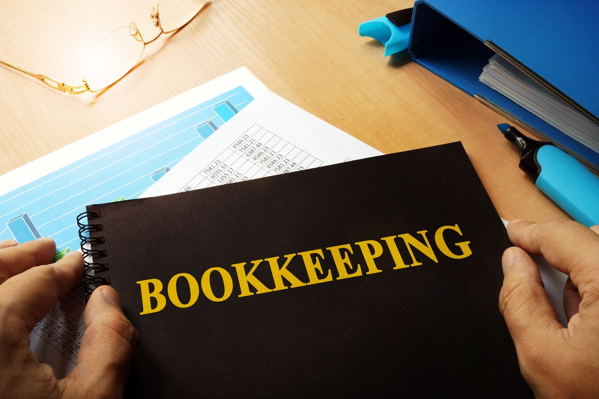Hands holding a black spiral-bound bookkeeping notebook on a desk, with other papers and a pen nearby.