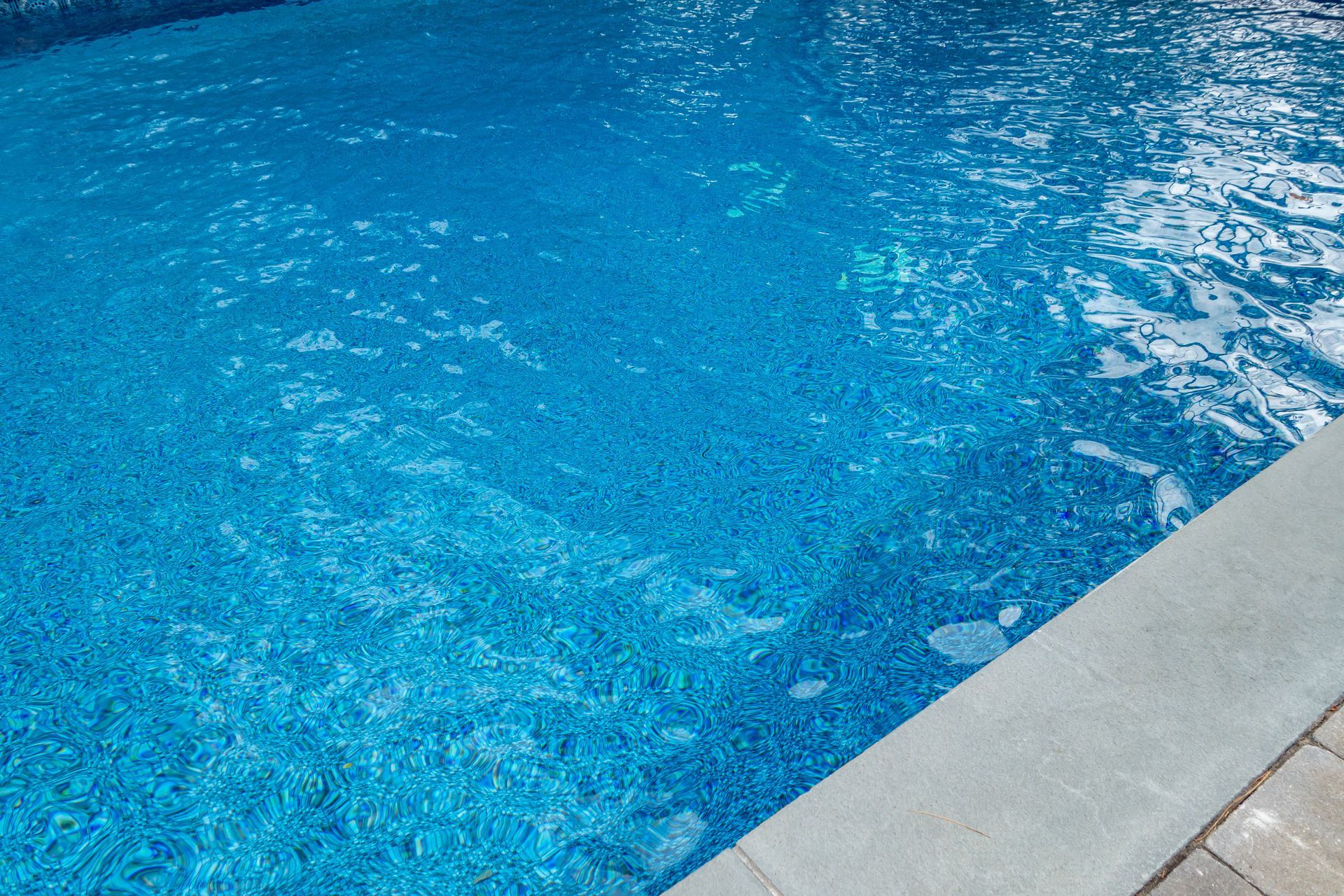 Close-up of clear blue swimming pool water with ripples beside a concrete edge.