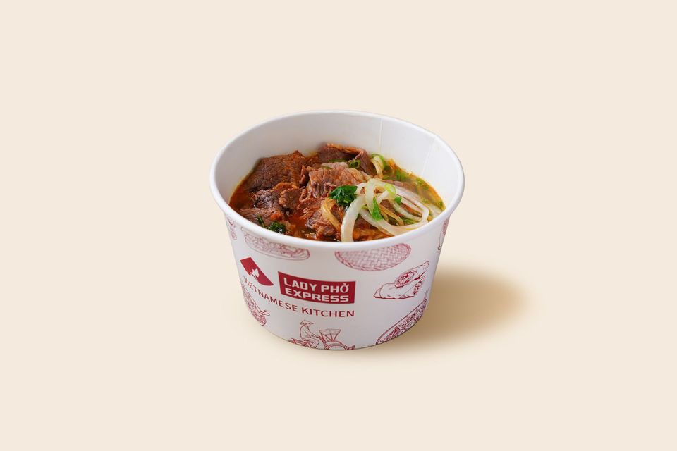 Cup of spicy noodles with green garnish in a white takeout container on a beige background