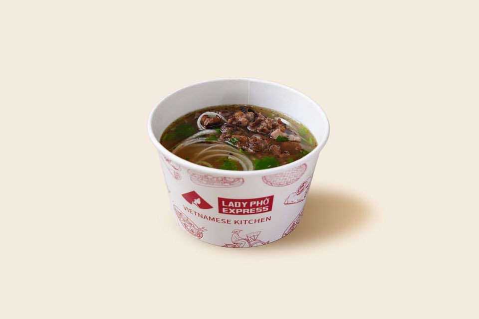 A paper cup of ramen soup with noodles and broth, set against a light beige background