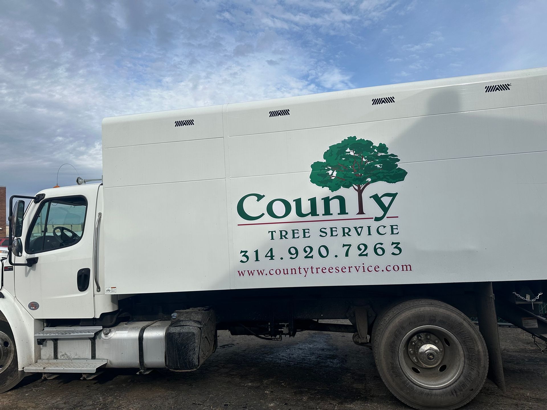 A side view of a white County Tree Service truck with a green tree logo, phone number, and website parked outdoors.