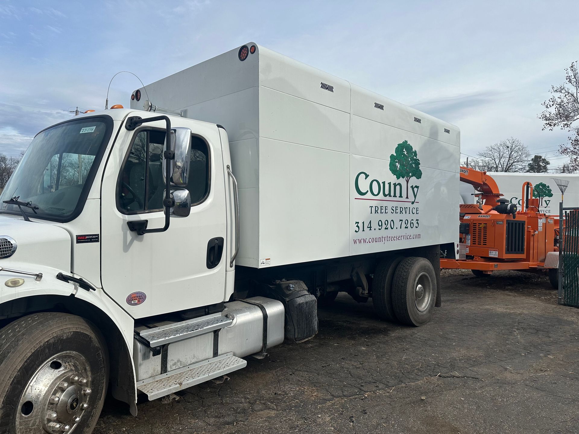 A white County Tree Service truck with an attached orange wood chipper parked on a gravel lot under a cloudy sky.
