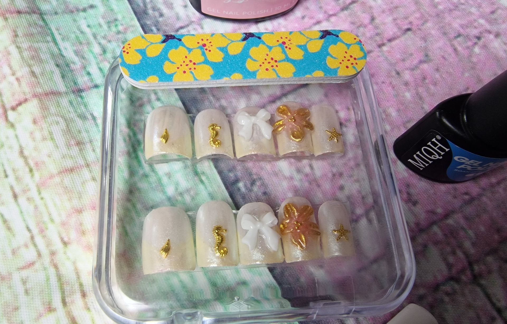 Diverse gouden nageltips in een transparant doosje op een pastelkleurige tafel.
