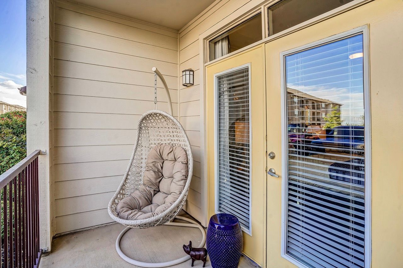 Balcony area with a white wicker hanging chair, beige walls, and glass double doors with blinds.