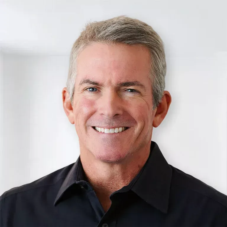Man with short gray hair, smiling, wearing a black shirt against a blurred white background.
