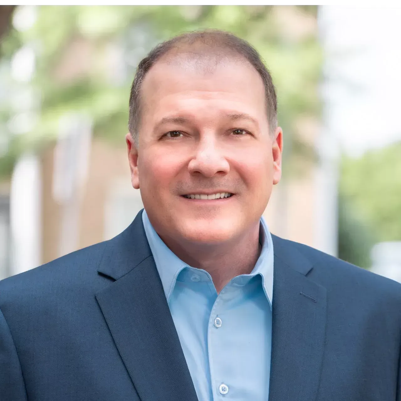 Man in blue blazer and shirt smiling outdoors.