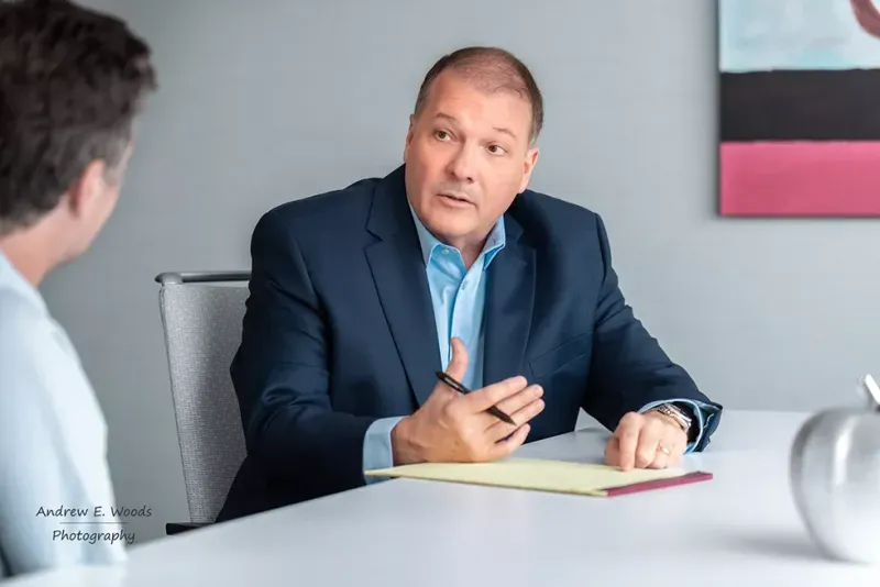 Man in suit gestures, speaking to another person at a table, likely in an office setting.