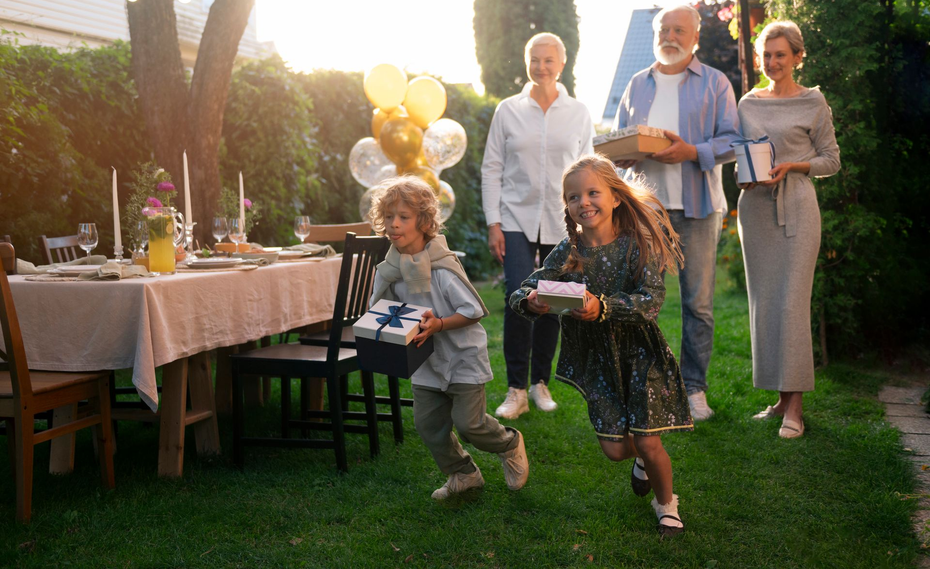 Children run with gifts toward a party table. Family follows on the lawn; balloons in background.