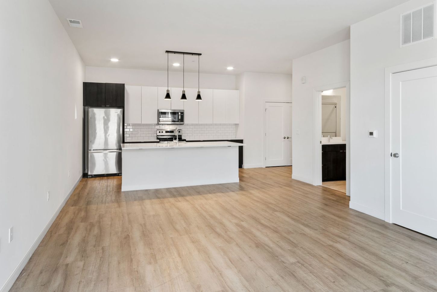 Modern open-concept living space with white walls, light wood floors, and kitchen with stainless steel appliances.