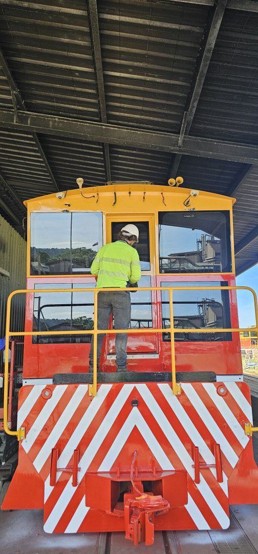 Front view of a red-and-yellow rail vehicle under a shed, with a worker standing on the rear platform