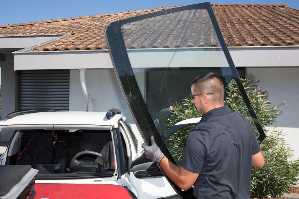 Man removing a broken windshield from a white car in a driveway by a house