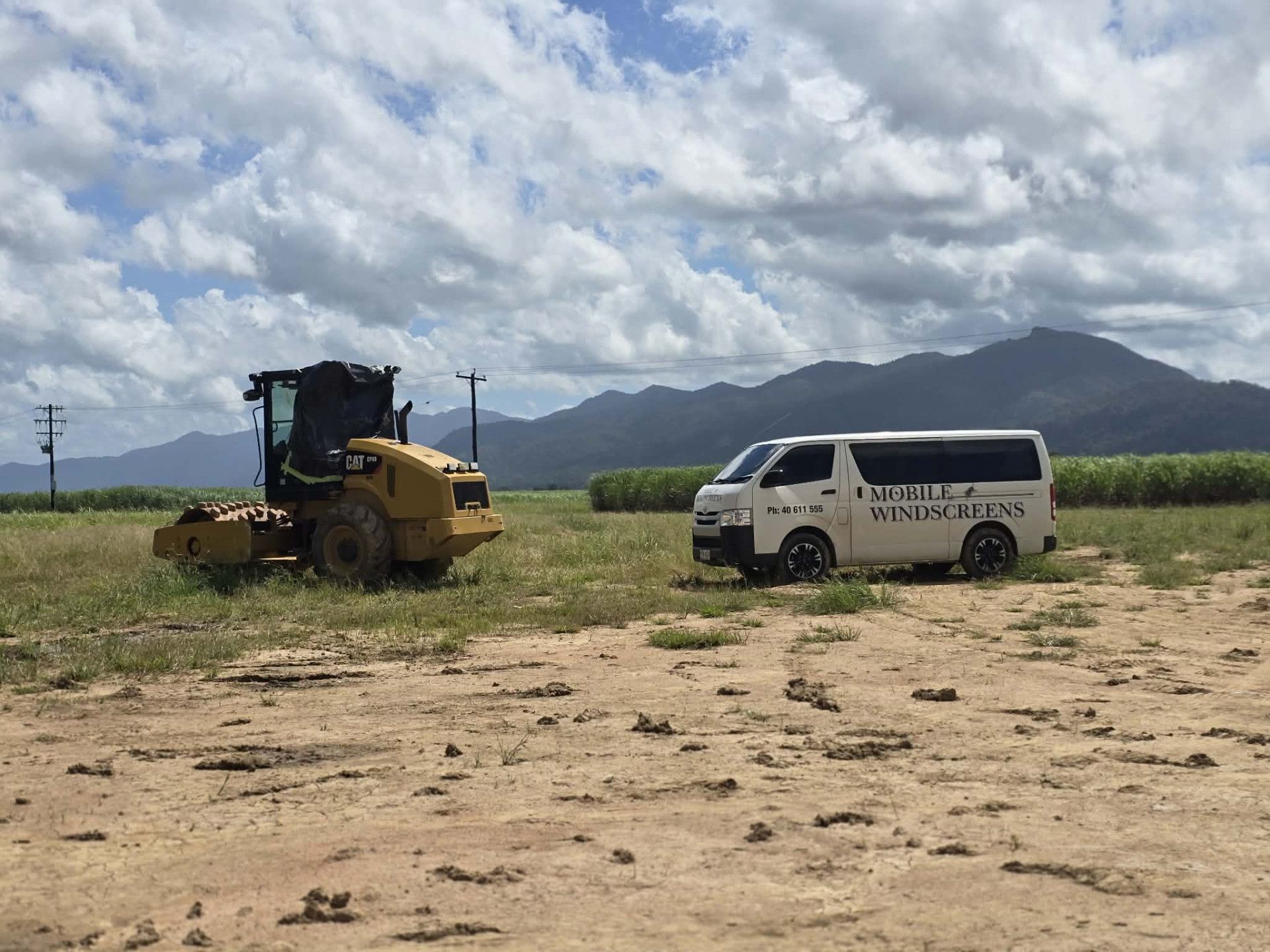 Yellow excavator and white van in a grassy field with mountains and cloudy sky in the background
