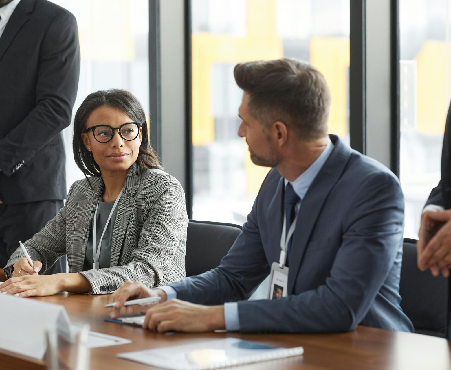 Two coworkers in suits talk at a conference table in a bright office.
