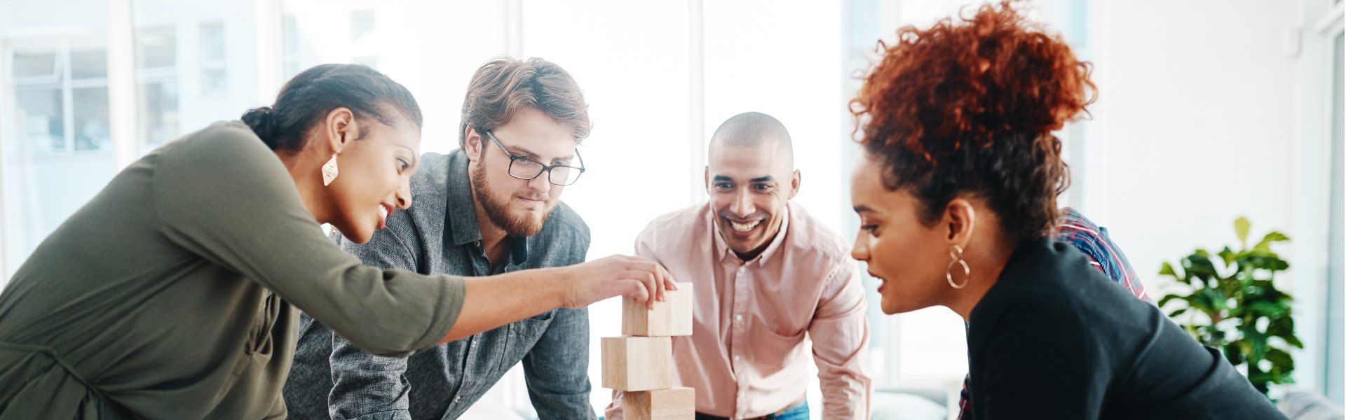 People collaborating around a tabletop tower-building game, reaching in and smiling in a bright room