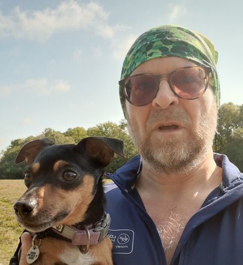 Man in sunglasses and bandana holds small dog outside.