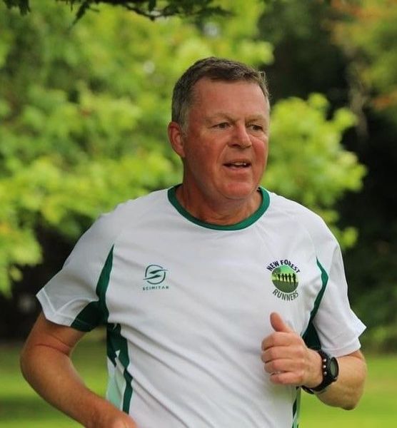 Man running outdoors, wearing white and green shirt with logo. Green trees in the background.