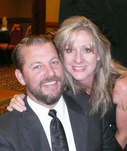 Smiling couple posing closely indoors, the man in a dark suit and tie, the woman in a dark sleeveless top.