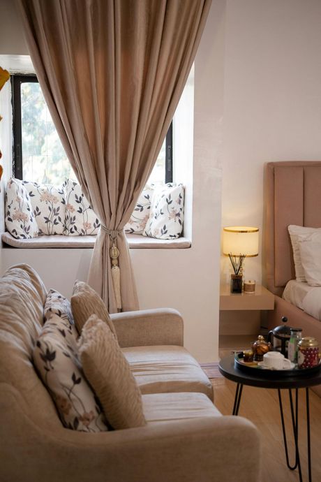 A beige sofa with floral pillows sits by a window with sheer curtains, next to a small side table and a bedroom area.