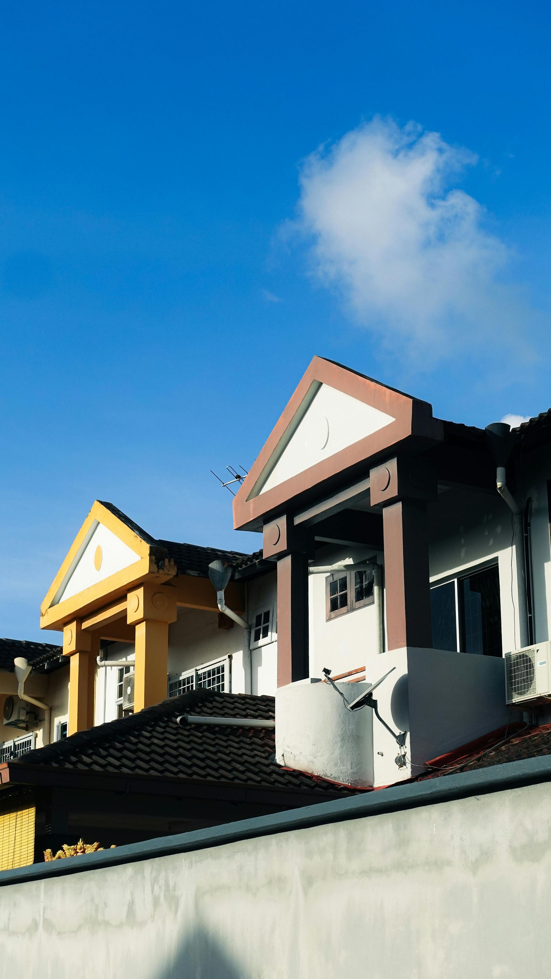 Two townhouses with triangular roof gables, one yellow and one brown, set against a bright blue sky.