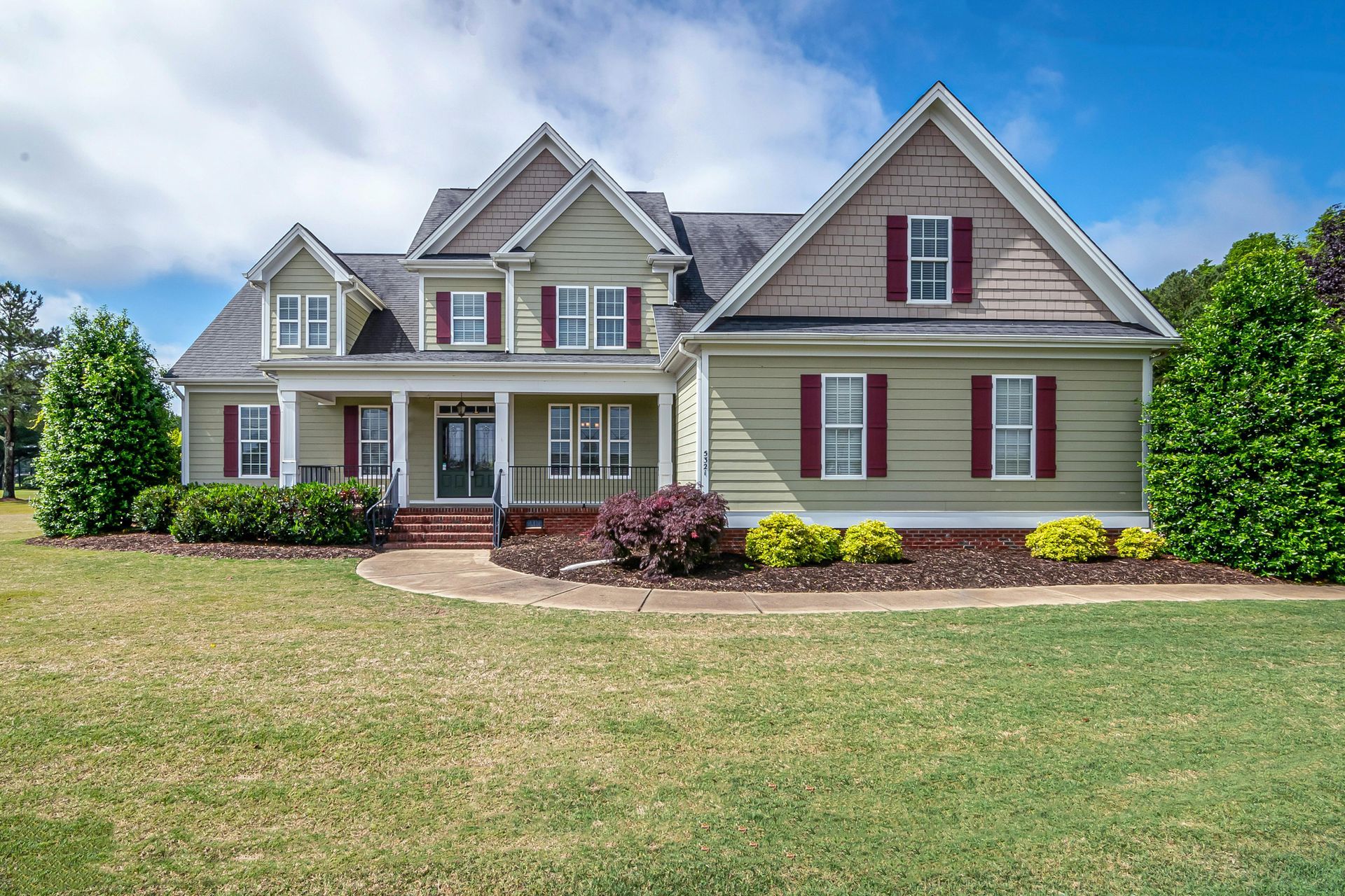 A two-story suburban house with light green siding, dark red shutters, a front porch, and a grassy yard under a blue sky.