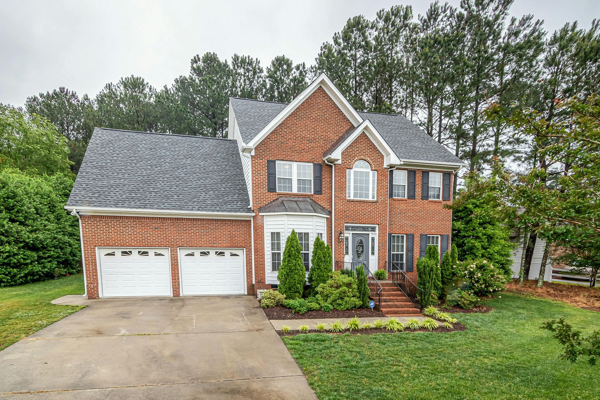 A two-story brick suburban house with a two-car garage, grey roof, and front lawn surrounded by green trees.
