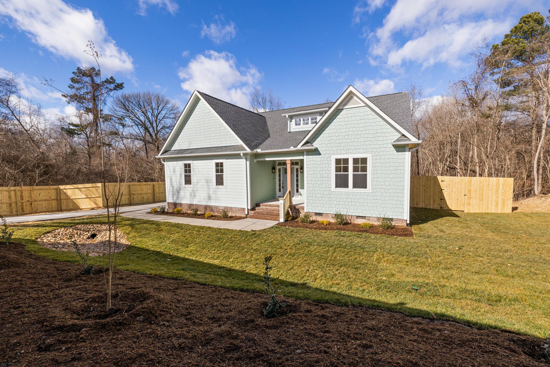 A light green house with a dark gray shingled roof and a wooden fence stands on a grassy lawn under a sunny blue sky.