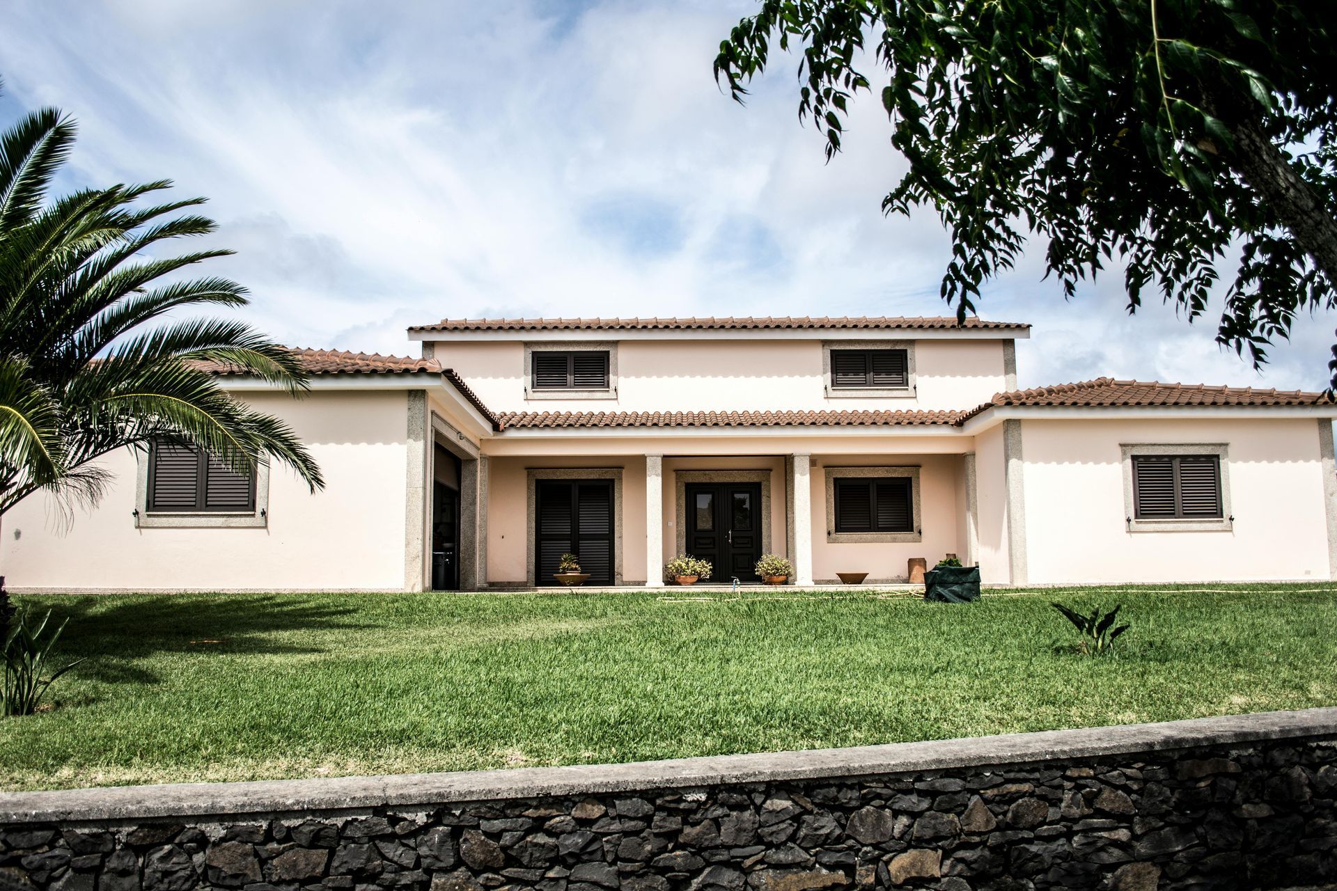 A pale peach-colored house with a dark stone base, brown window shutters, and a green lawn under a partly cloudy sky.