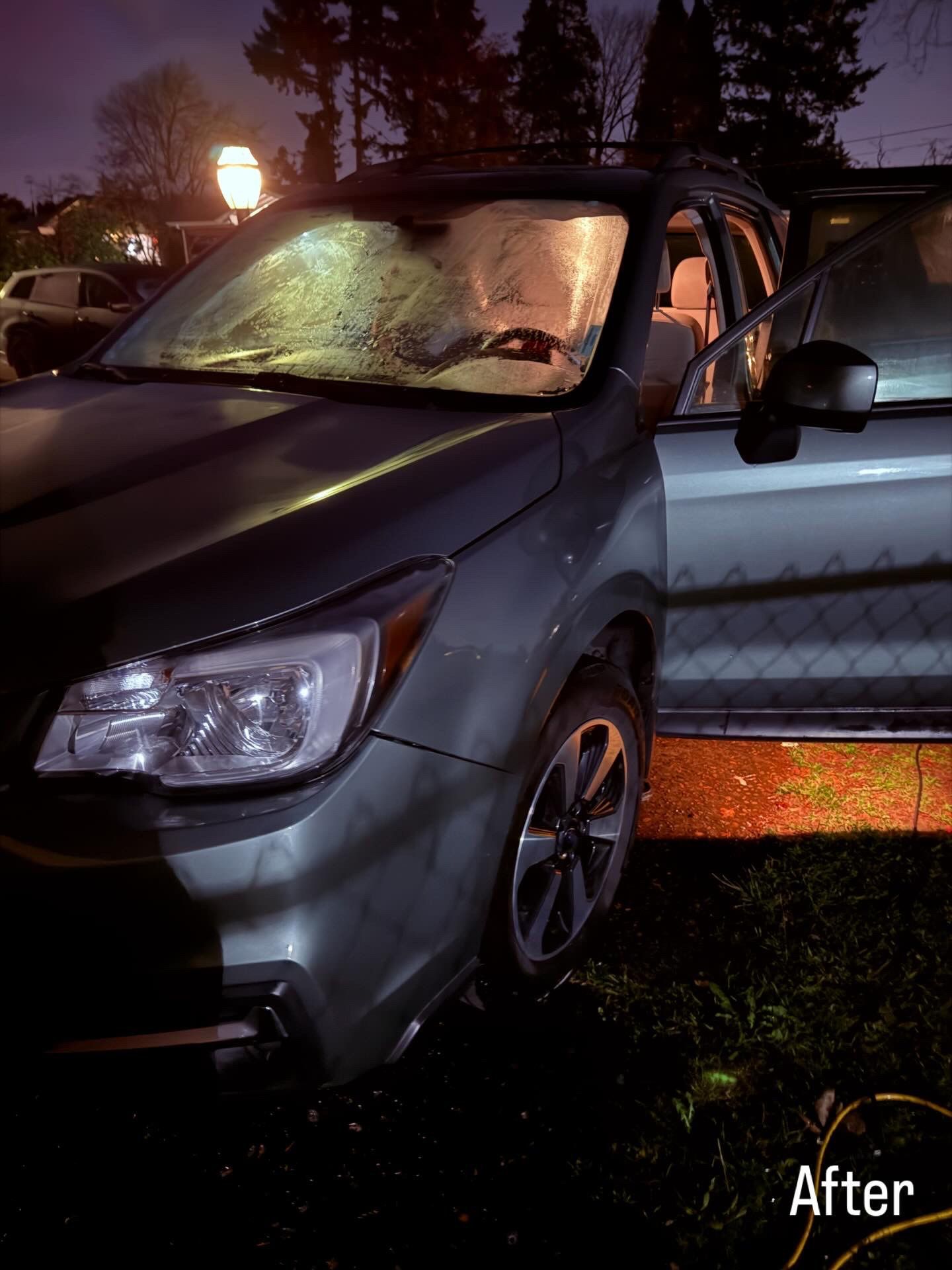 Front-left view of a dark sedan with a damaged windshield and open door at night, lit by streetlights.